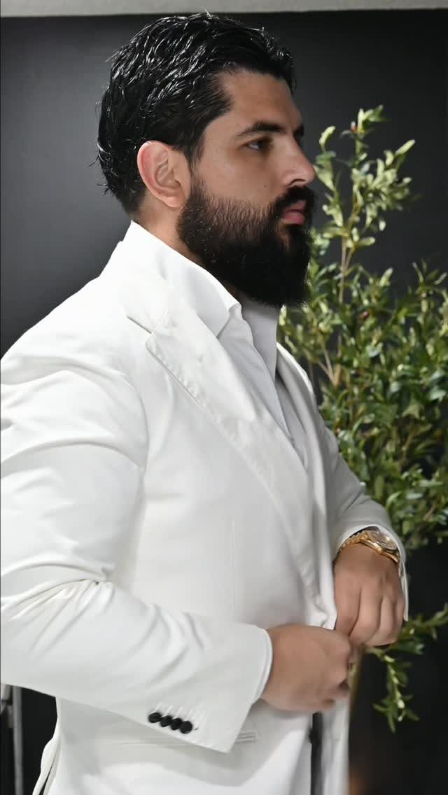A bearded man in a white suit jacket posing against a dark background with plants.
