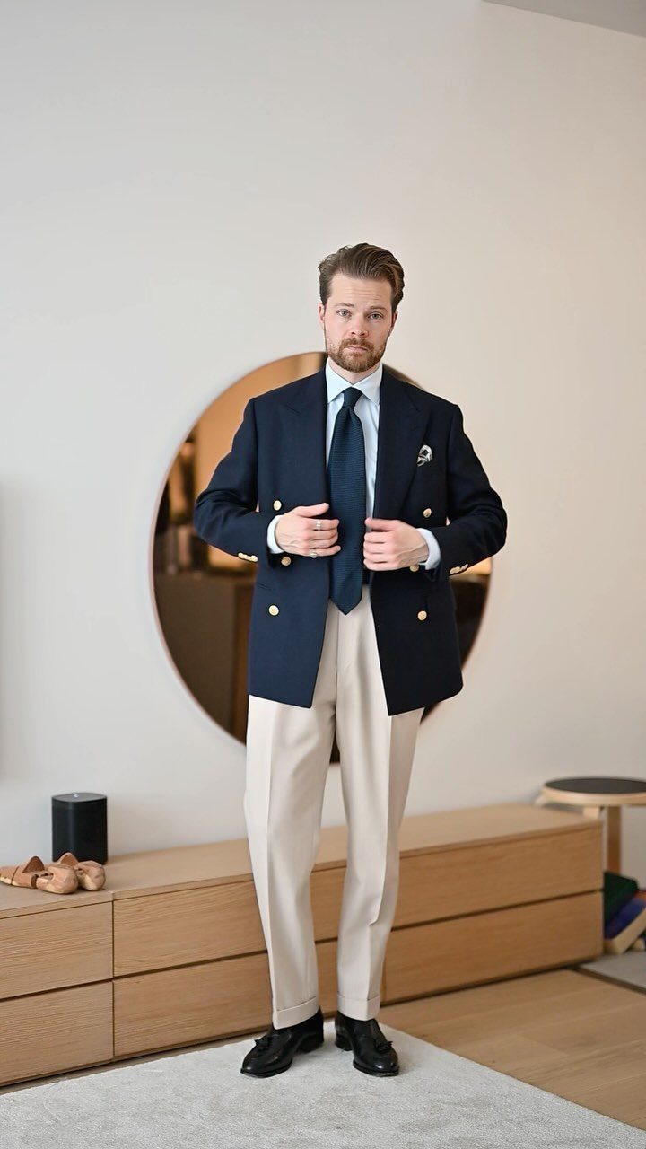 Man wearing a navy double-breasted blazer, light blue shirt, navy tie, and beige trousers.
