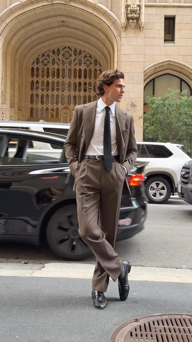 Man in a brown tailored suit, white shirt, and black tie standing on a city street.