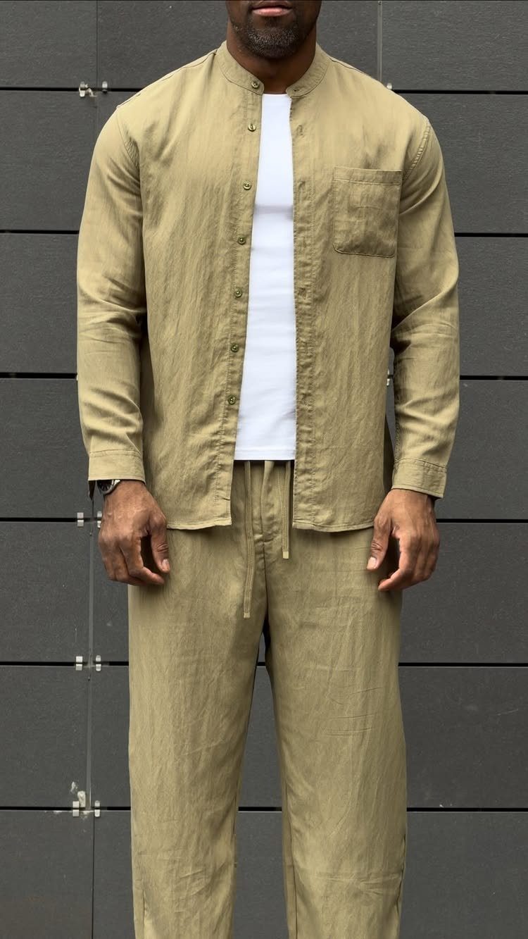 Man wearing a tan linen shirt and matching linen drawstring trousers against a dark wall.