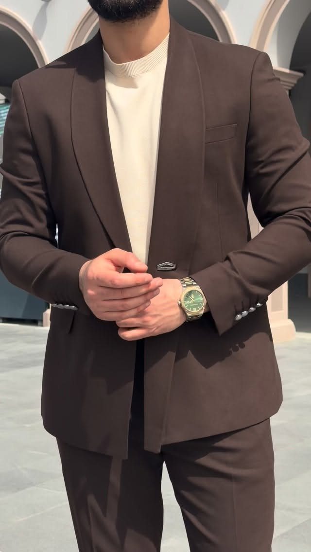 Man in a dark brown tailored suit and cream sweater standing outdoors.