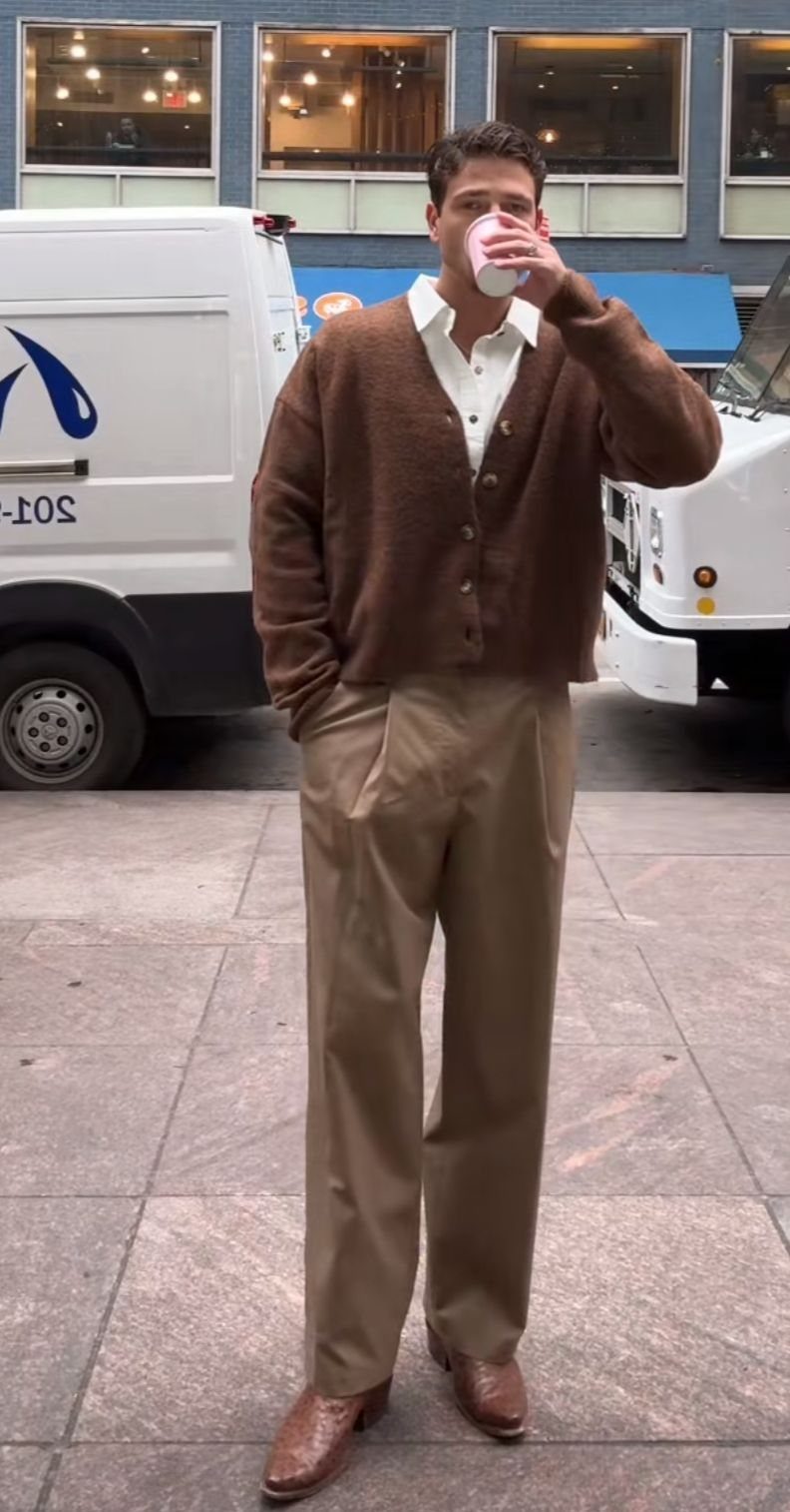 Man in a brown cardigan and tan trousers drinking from a pink cup on a city street.