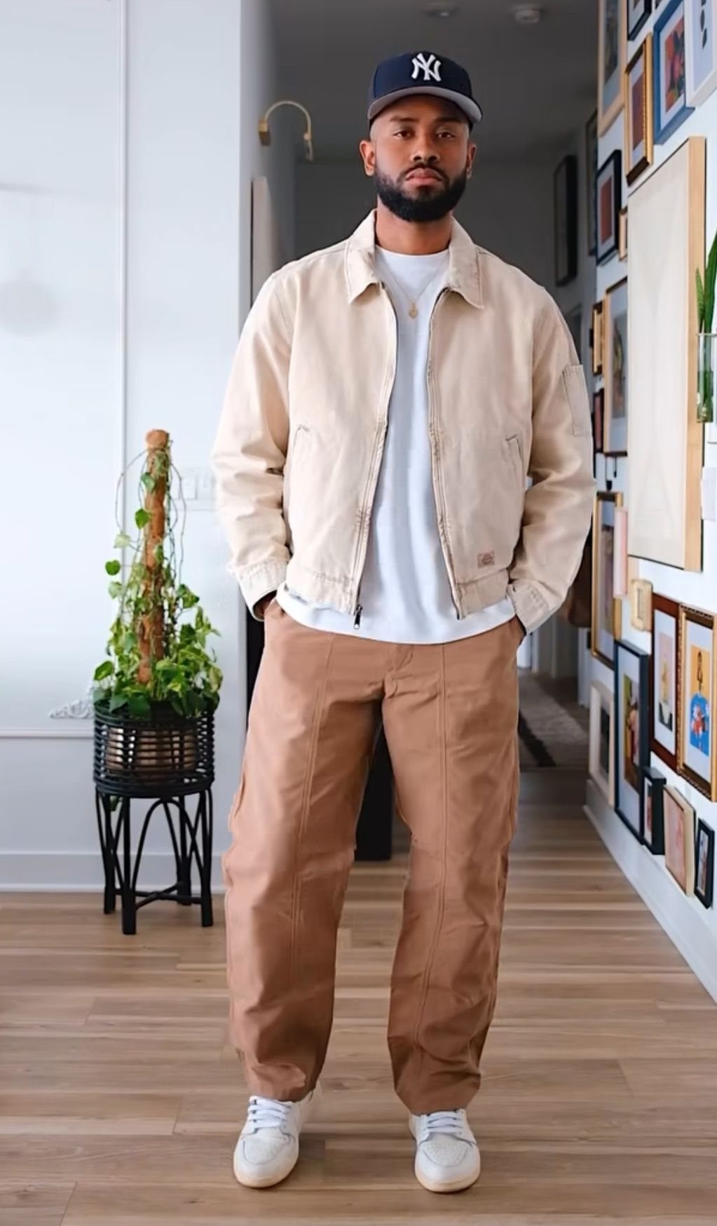 A man wearing a cream jacket, white t-shirt, tan pants, and a navy baseball cap.