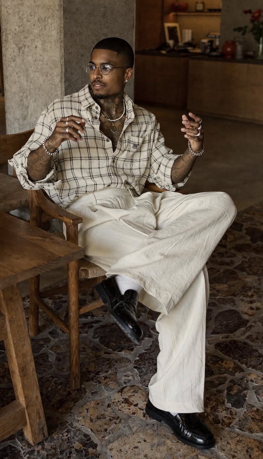 A man wearing a plaid shirt and beige trousers sitting in a wooden chair.