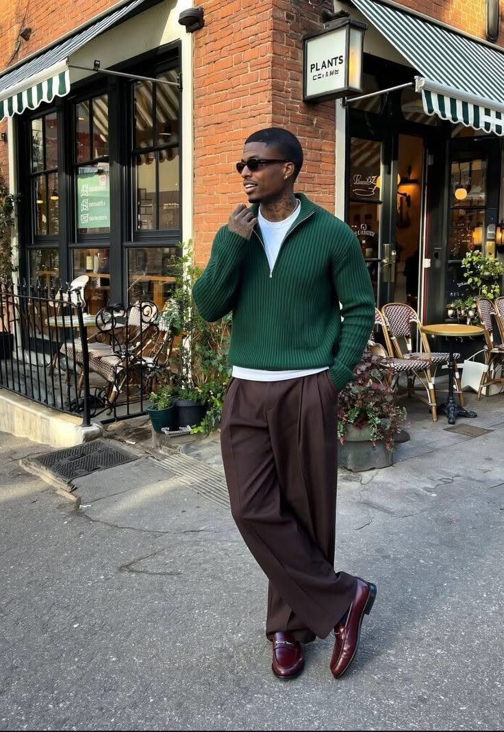 Man wearing a green sweater, brown trousers, and burgundy loafers standing outside a brick cafe.