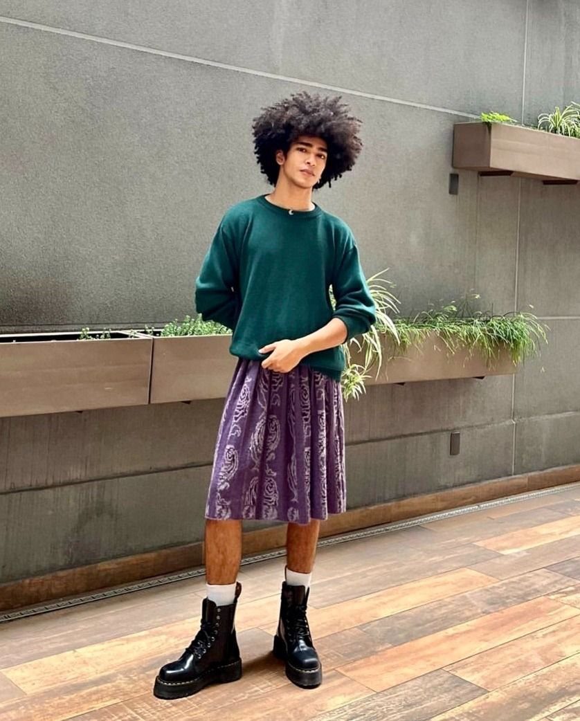 A person wearing a green sweater and a purple patterned skirt with black boots.