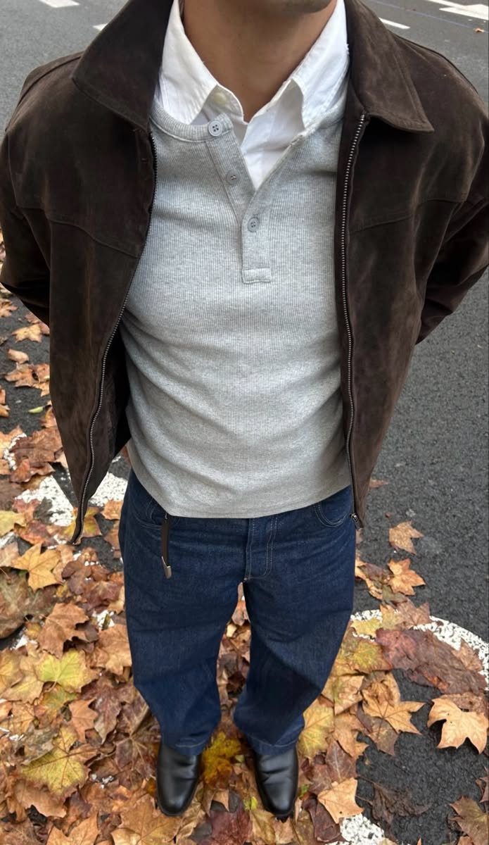 A person wearing a brown suede jacket, grey polo, and blue jeans standing among autumn leaves.