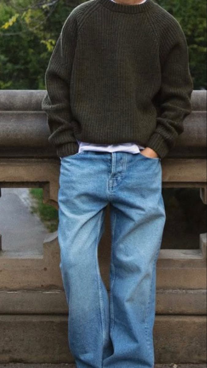A person wearing an olive green ribbed sweater and wide-leg blue jeans standing by a stone railing.