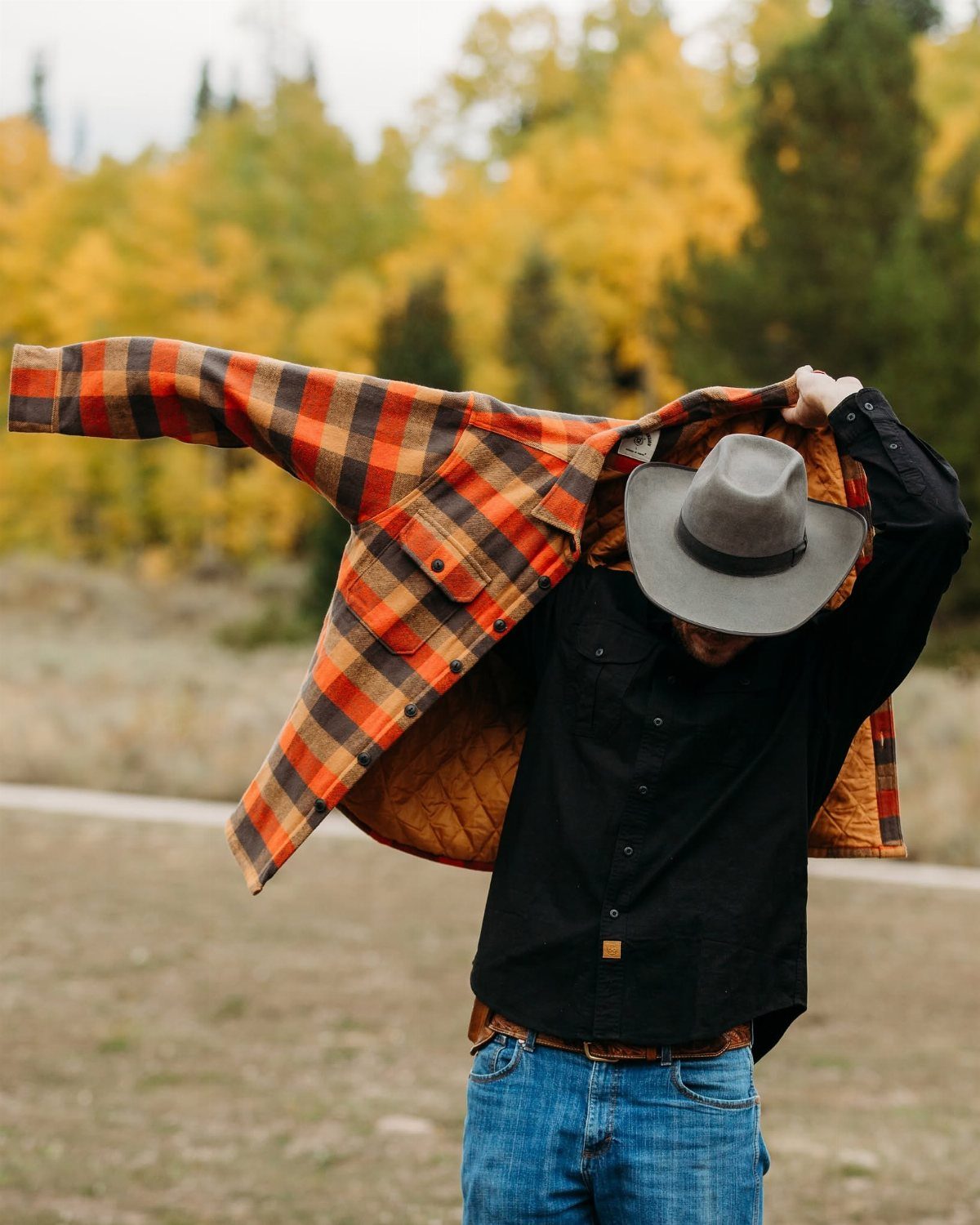 Person in a black long-sleeved button-up shirt and cowboy hat holding an orange plaid jacket.