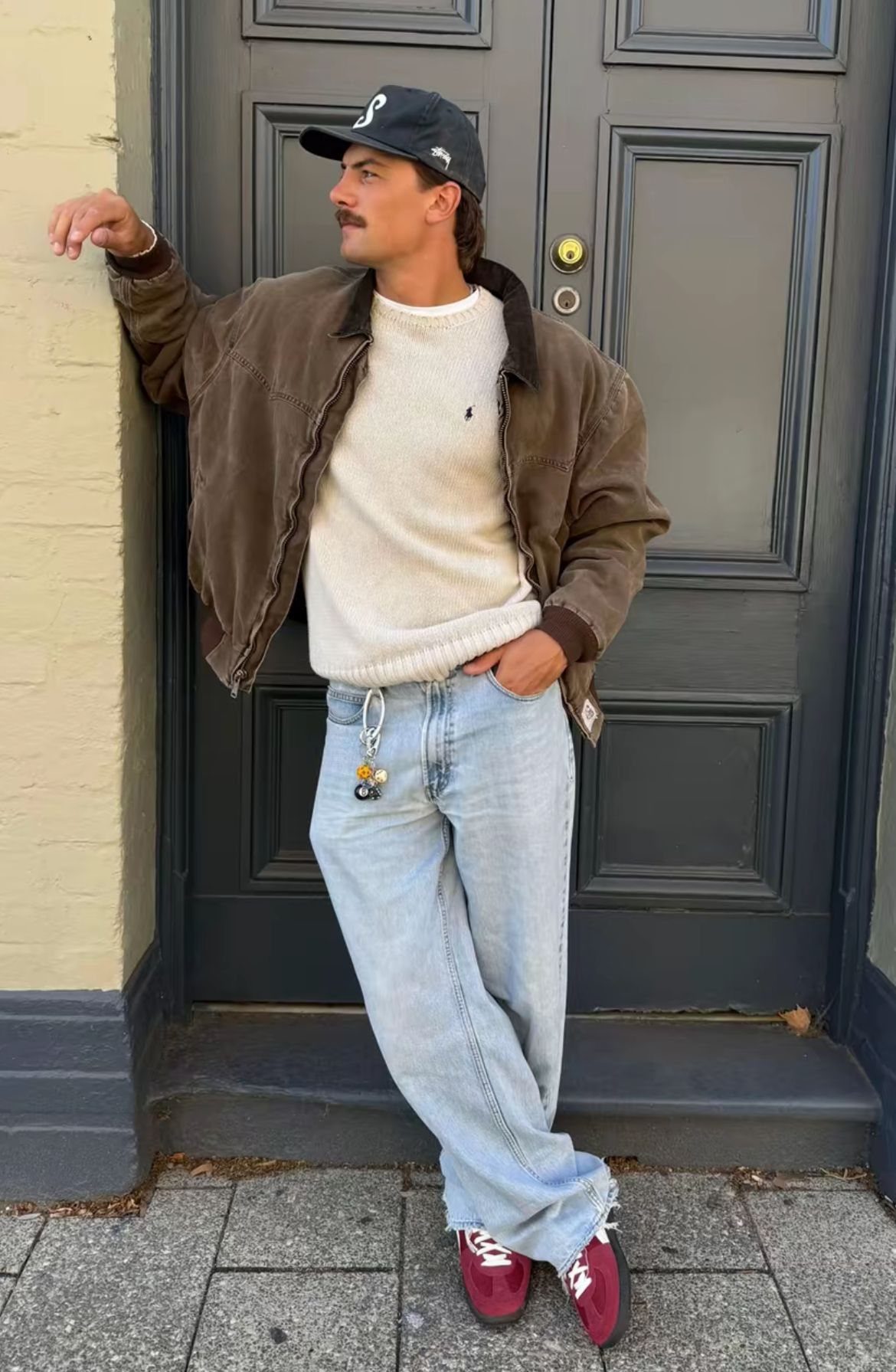 Man in a cream sweater and brown jacket wearing light blue low-slung jeans.