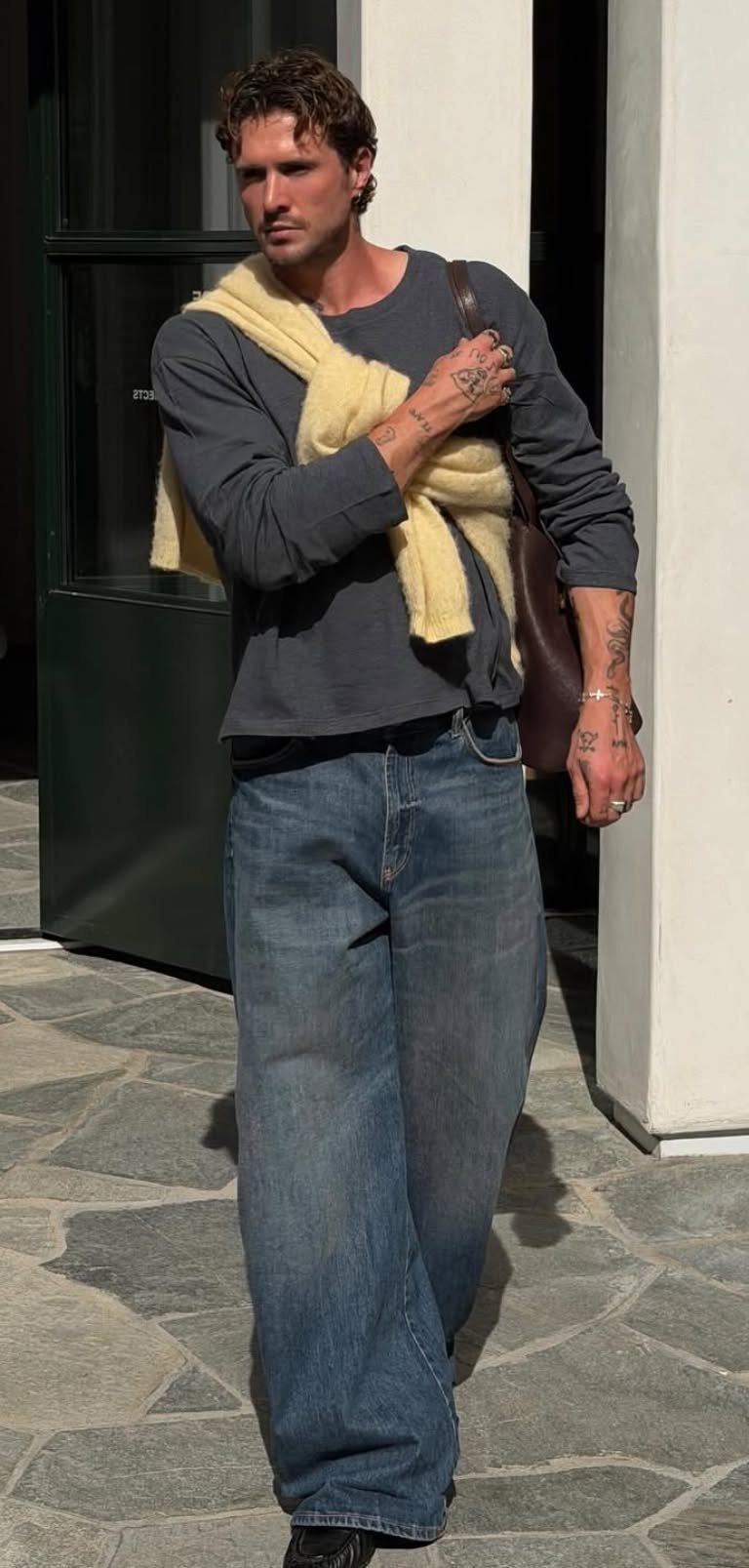 A man walking in a grey long-sleeve shirt, wide blue jeans, and a yellow sweater draped over his shoulders.