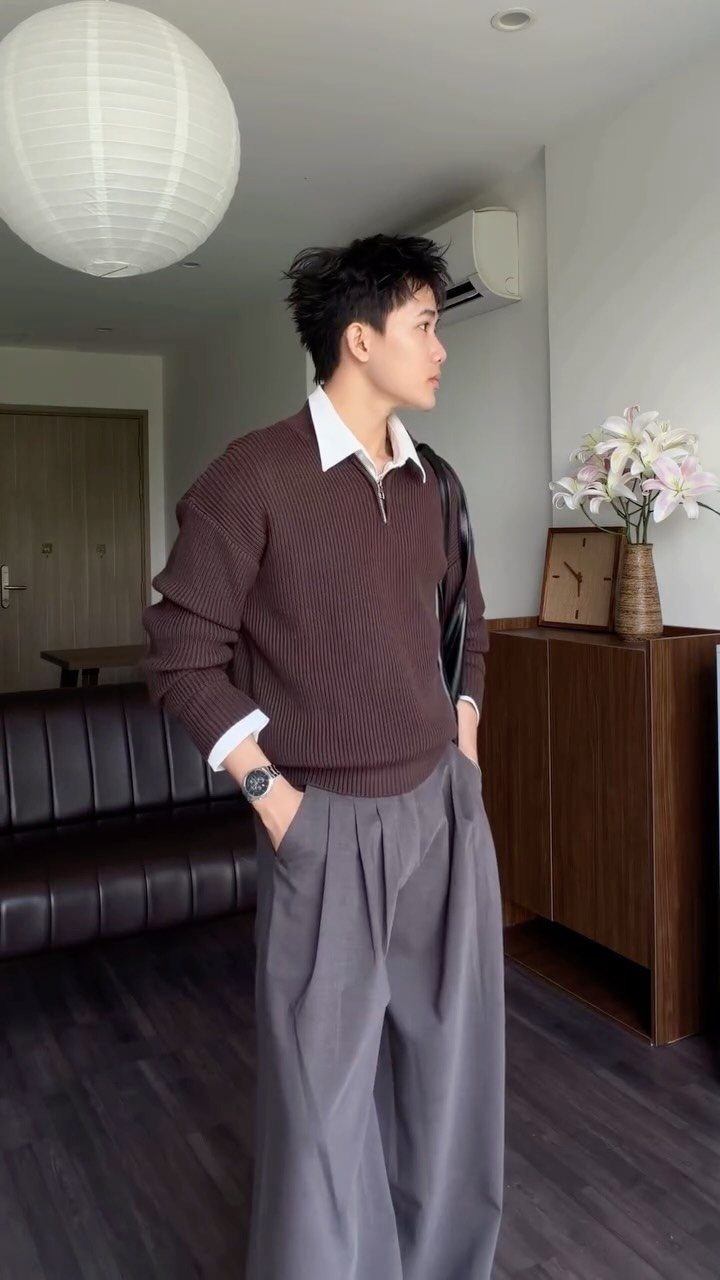A man wearing a brown ribbed sweater and wide-leg grey pleated trousers standing indoors.