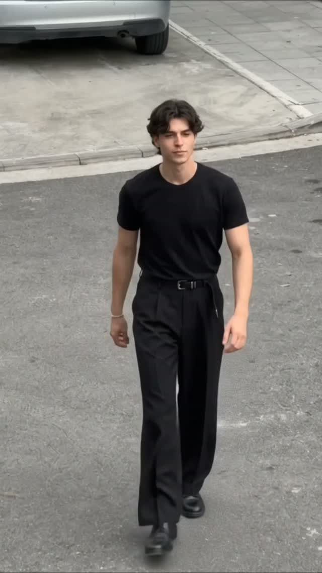 A man walking down a street wearing a black t-shirt and wide-leg black pants.