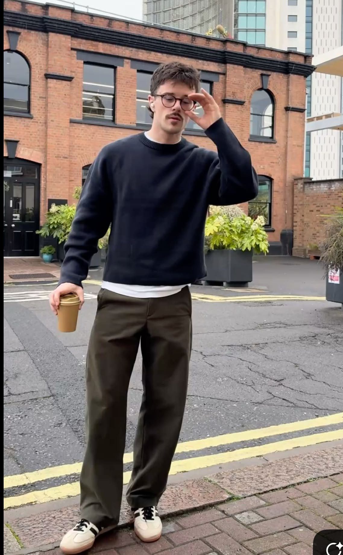 Man in navy sweatshirt and olive trousers standing on a city street holding a coffee cup.