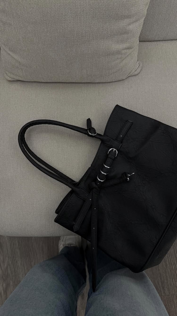 A black leather tote bag resting on a grey sofa next to blue jeans.