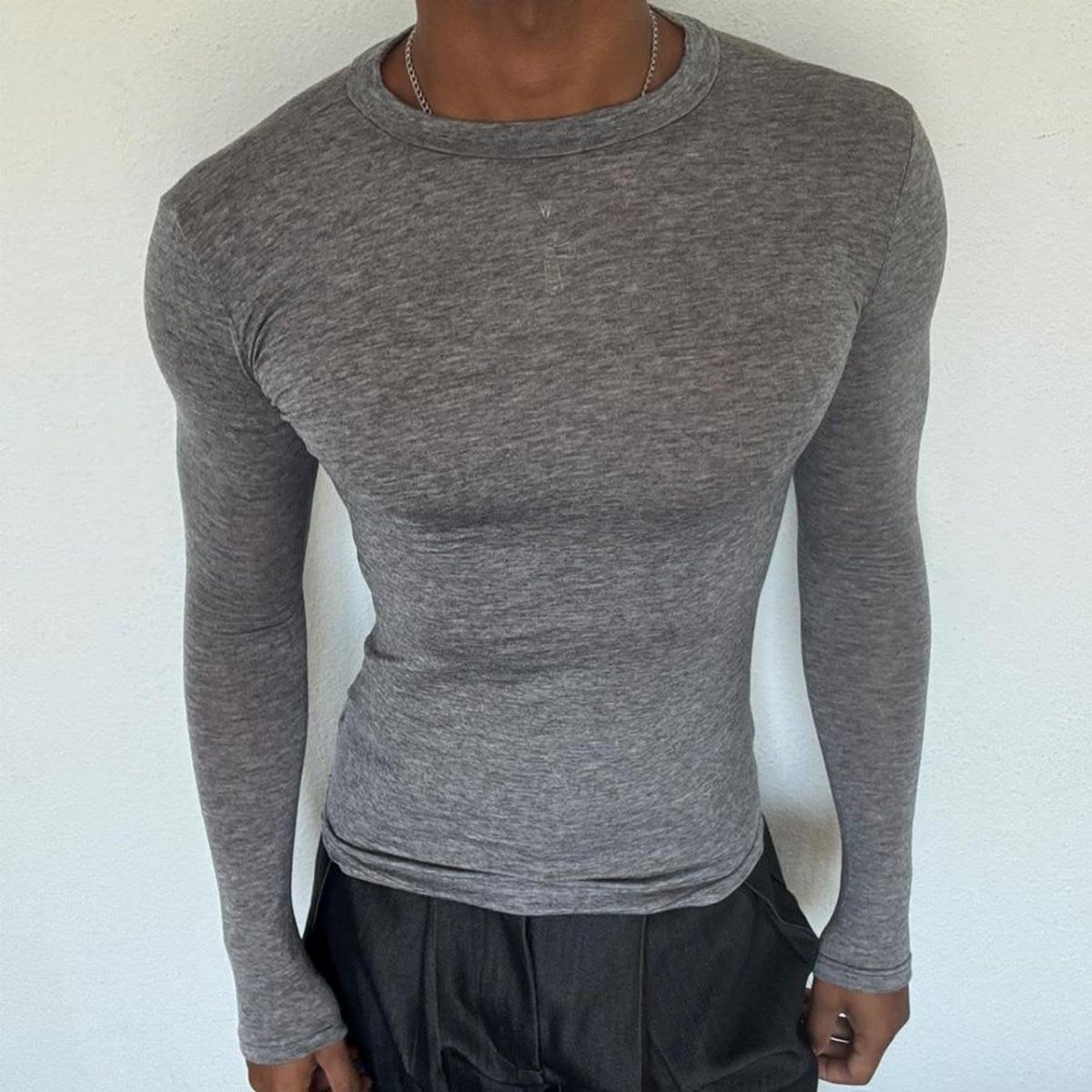 A person wearing a fitted grey long sleeve shirt and black pants against a white wall.