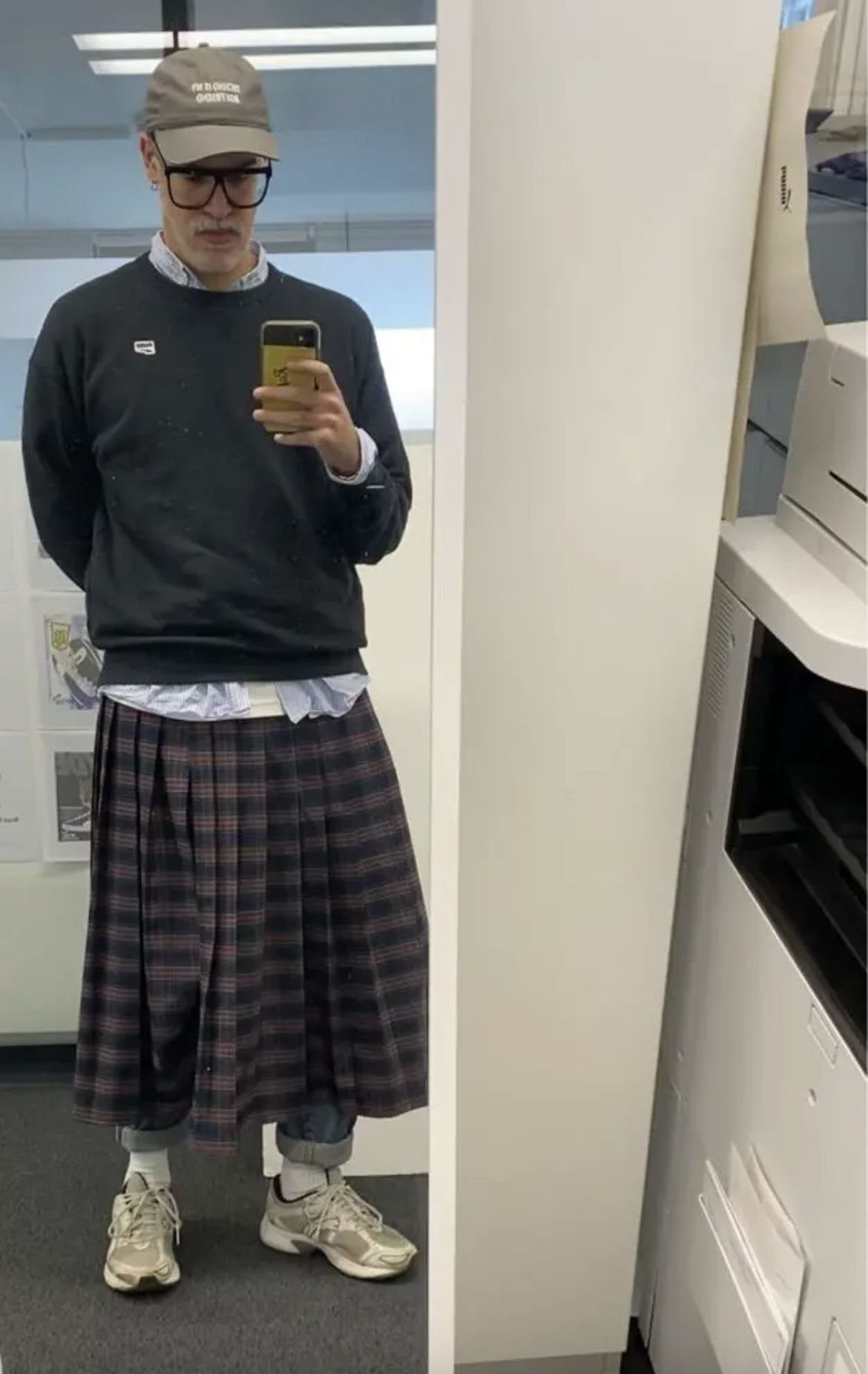 A person wearing a black sweatshirt, plaid skirt, tan cap, and sneakers taking a mirror selfie.