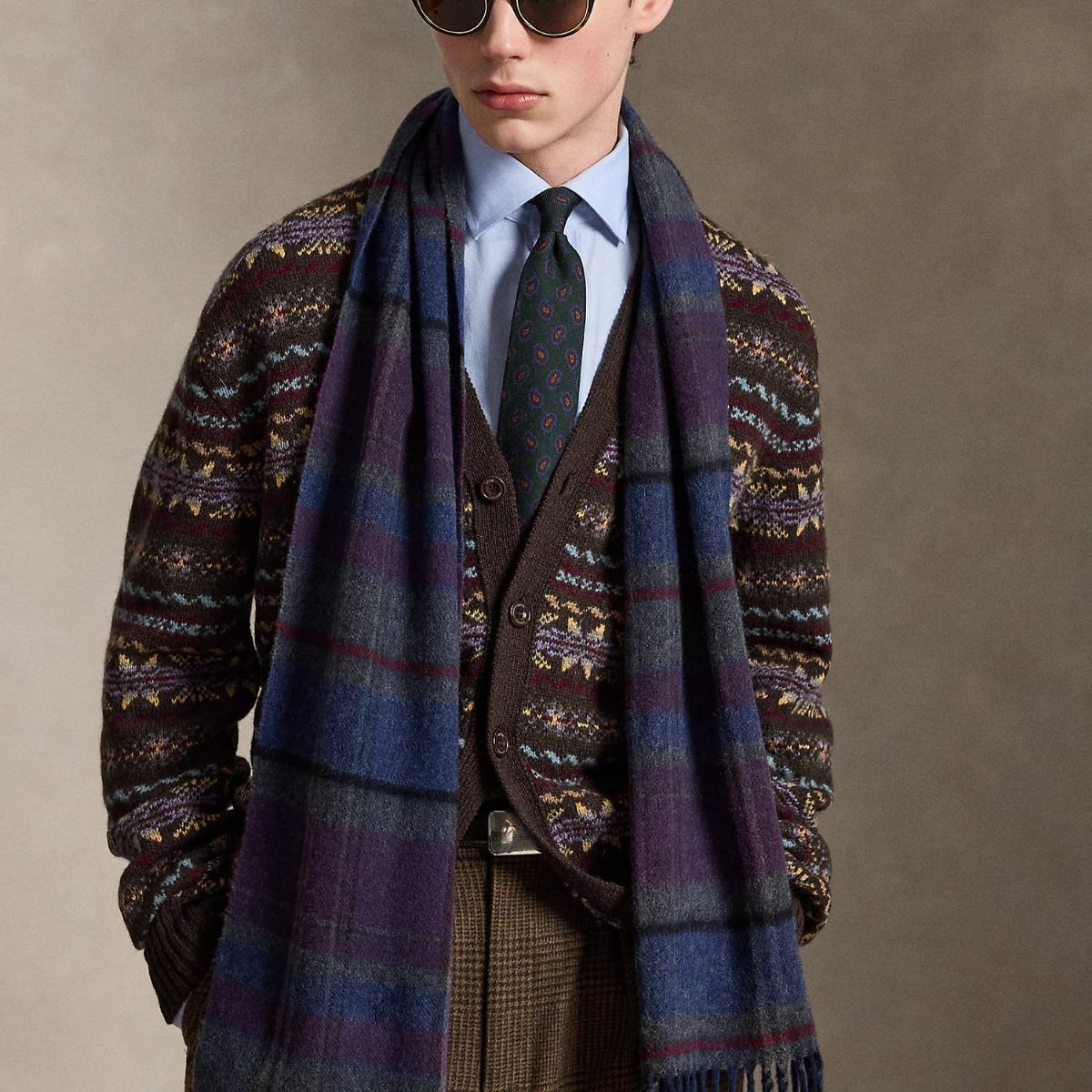A man wearing a patterned cardigan and a large blue and purple plaid scarf.