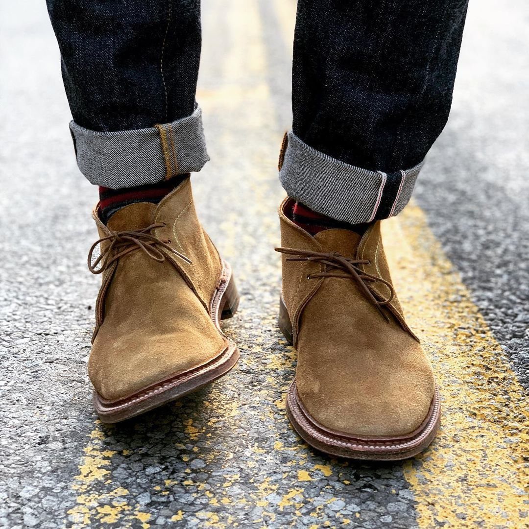 Person wearing dark denim jeans and tan suede boots walking on an asphalt road.