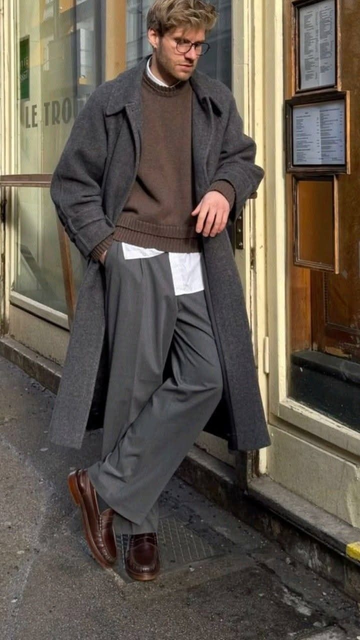 A man wearing a long grey coat, brown sweater, and grey trousers leaning against a building.