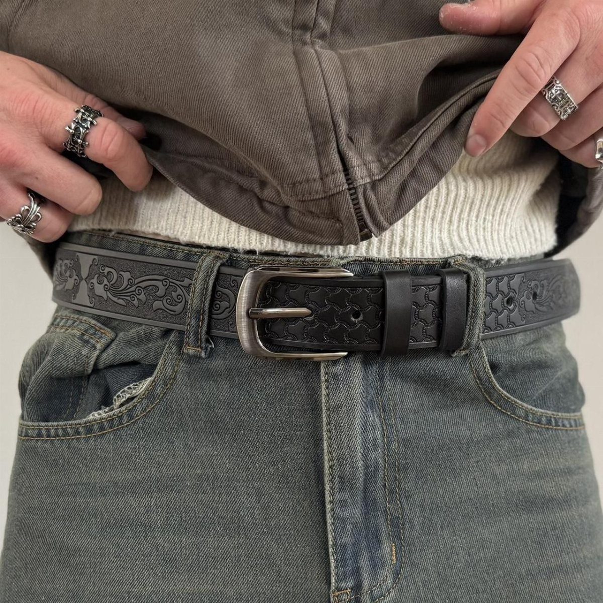 A person wearing a patterned black leather belt, silver rings, and blue denim jeans.