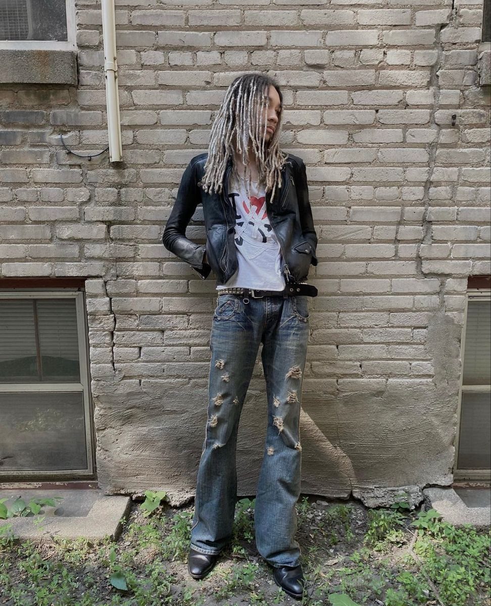 A person with dreadlocks wearing a leather jacket, graphic t-shirt, and distressed jeans against a brick wall.