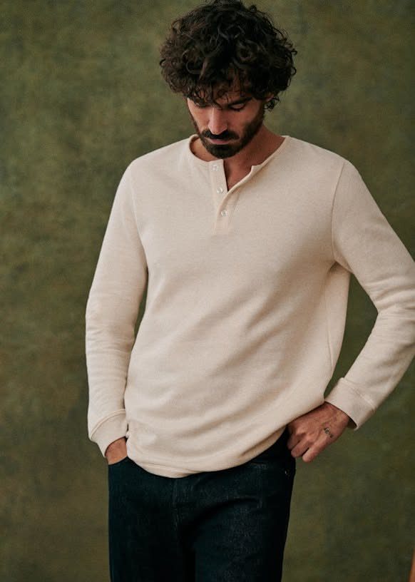 A man with curly hair wearing a cream long-sleeve henley shirt and dark pants.