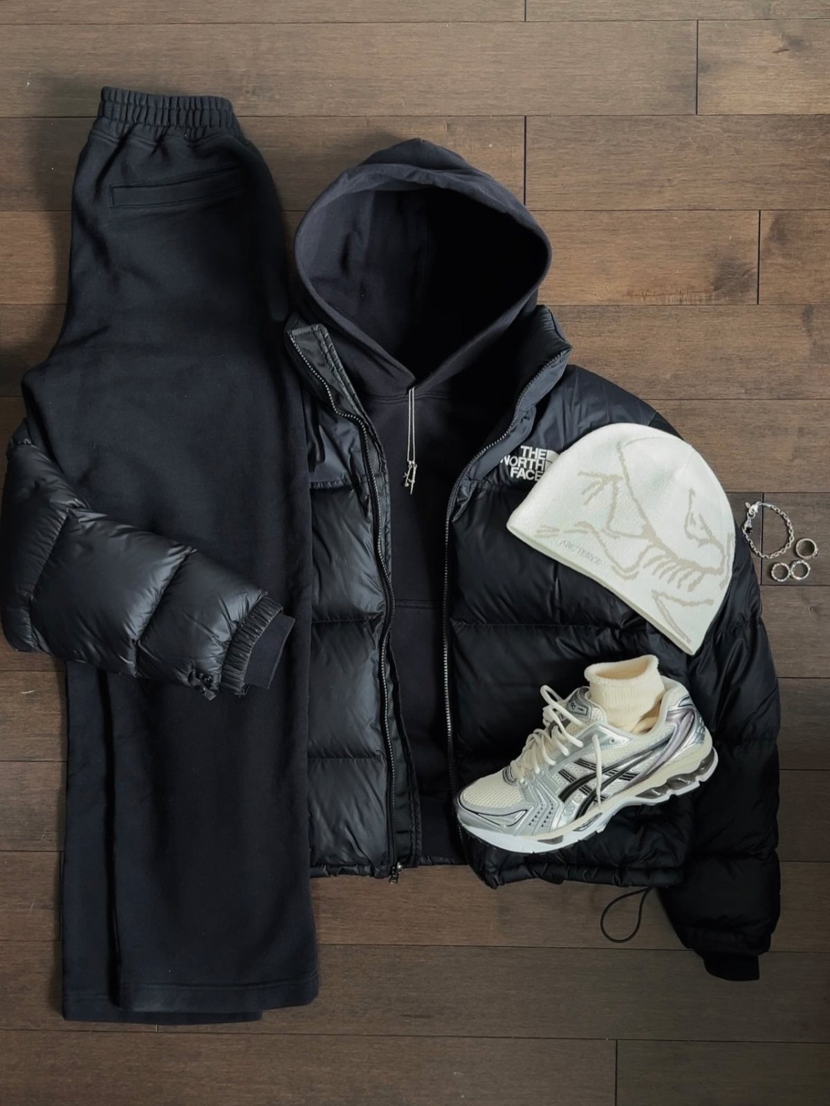 Black puffer jacket, hoodie, pants, white beanie, and sneakers laid out on a wooden floor.