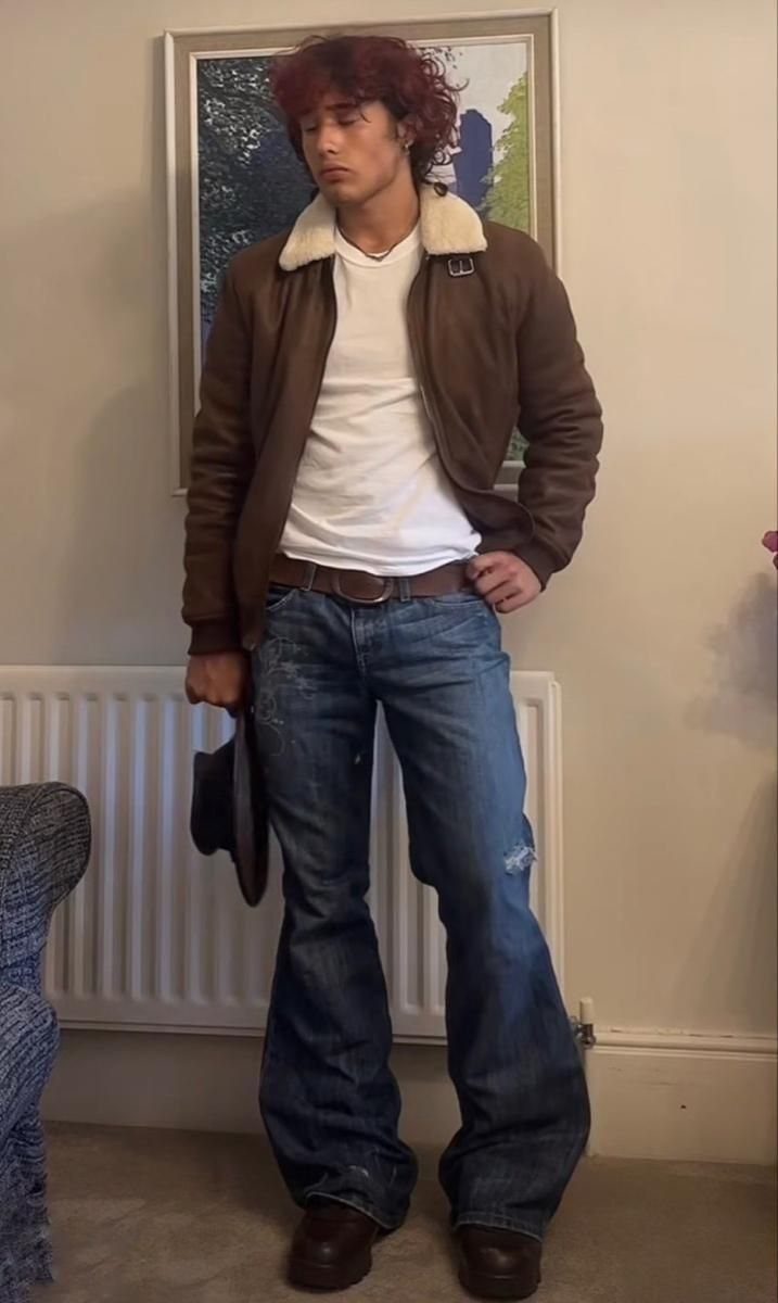 A young man posing in a brown bomber jacket, white t-shirt, and blue flared jeans.