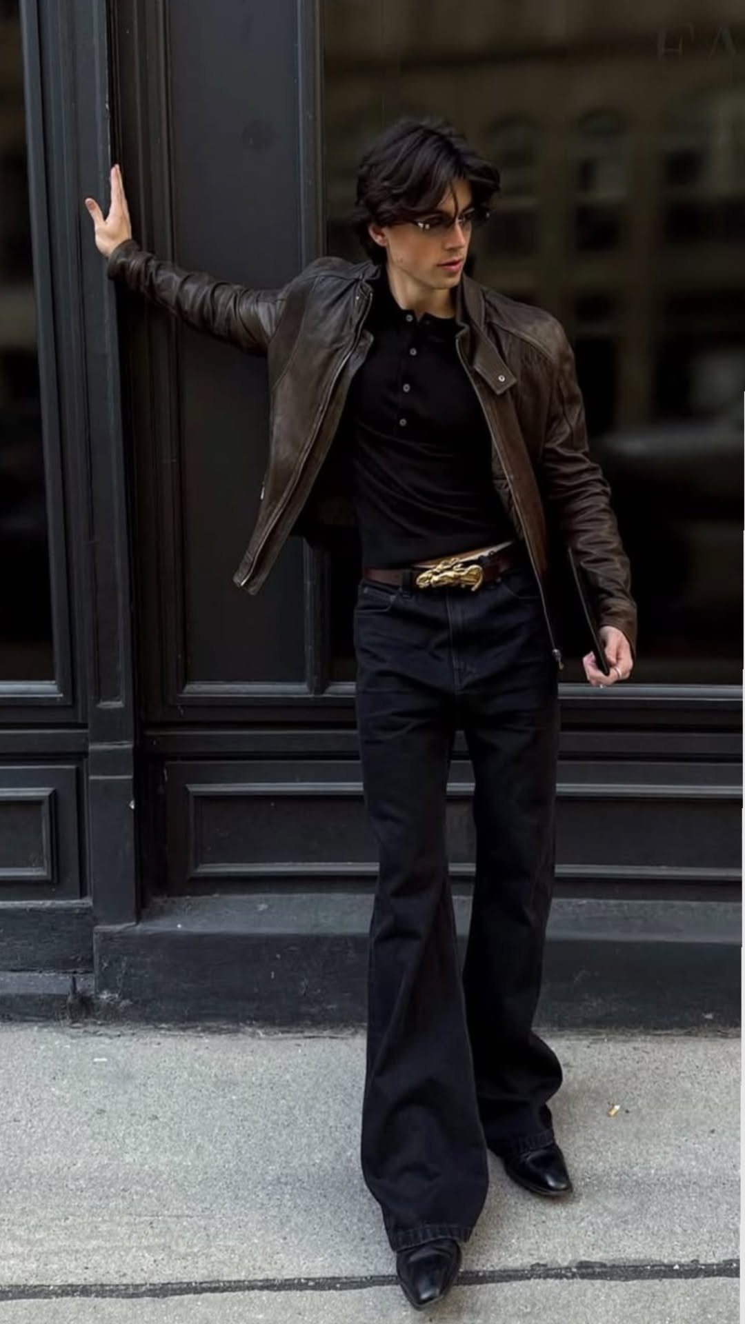 A person in a leather jacket and black flare trousers posing against a dark building.