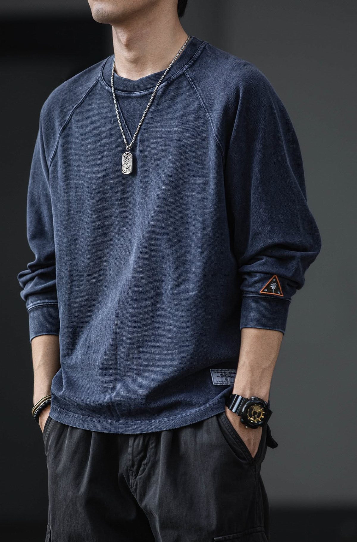 Man wearing a faded blue long-sleeved sweatshirt and black trousers with a silver necklace.