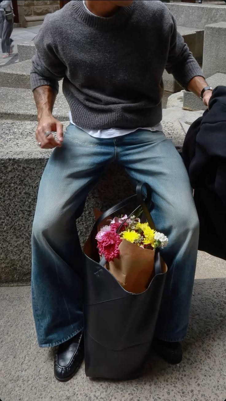 A person in a grey sweater and blue jeans sits with a flower-filled tote bag.