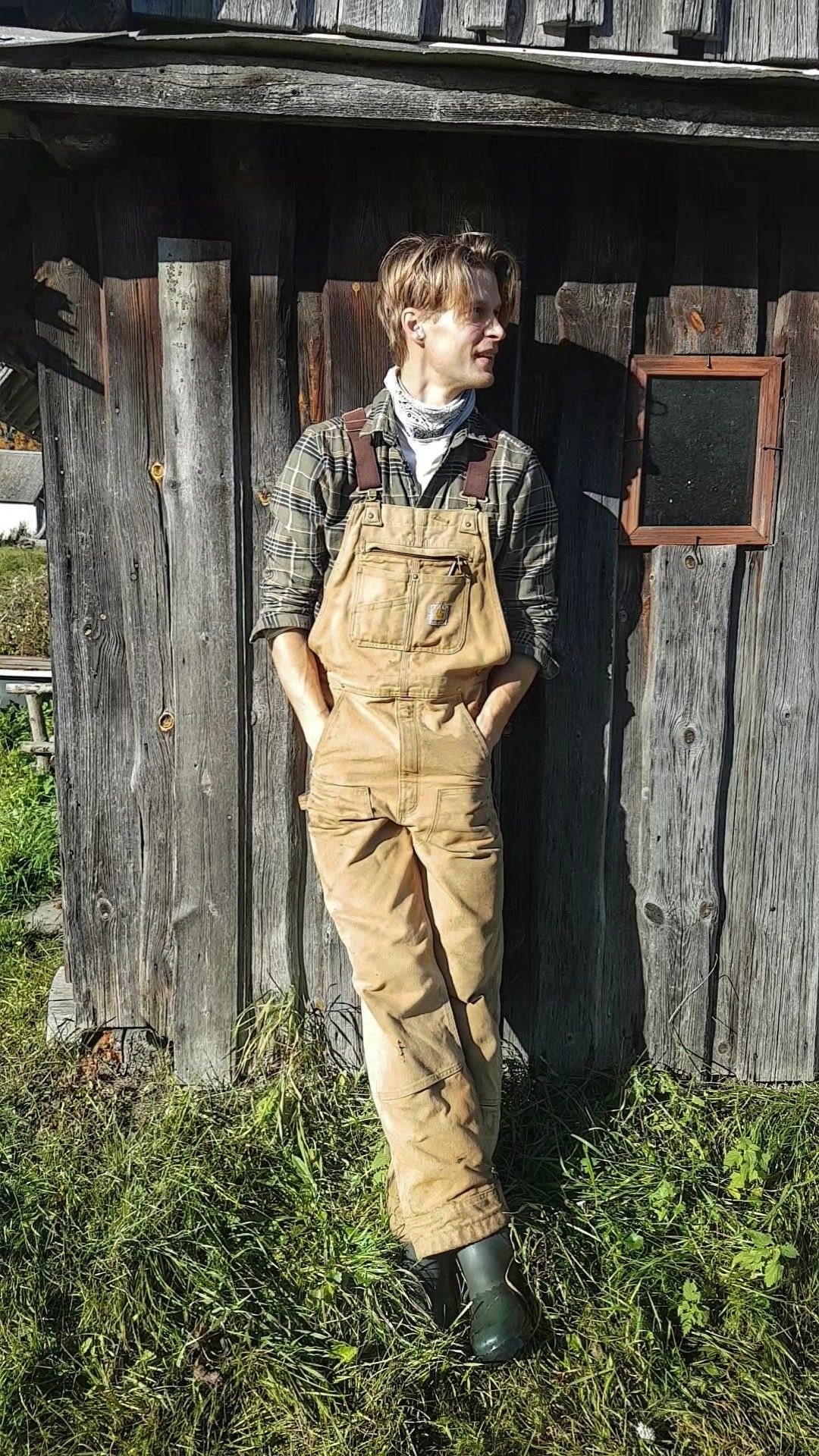 A young man posing in tan overalls and a plaid shirt against a wooden wall.