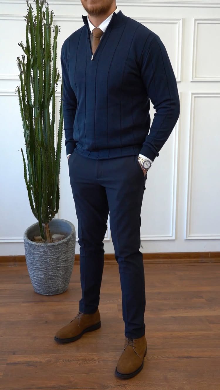 Man wearing a navy blue zip-up sweater, white shirt, tie, and navy trousers standing near a cactus.