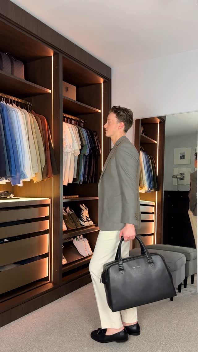 A man in a grey blazer and white trousers holding a black leather bag.