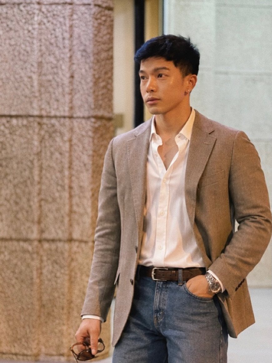 A man wearing a tan blazer, white shirt, and blue jeans standing outdoors.