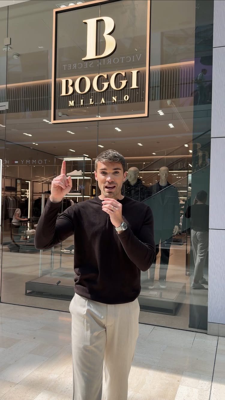 Man wearing a dark knit sweater and light trousers standing in front of a clothing store.