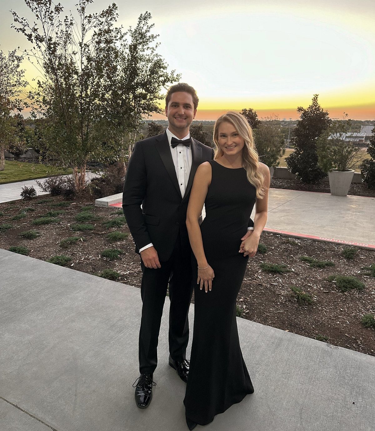 A man in a black tuxedo and a woman in a black dress posing outdoors at sunset.