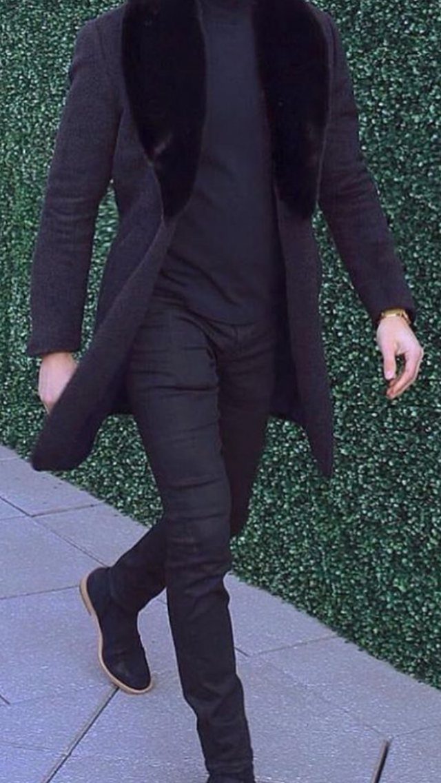 Man in an all-black winter outfit walking past a green hedge on a sidewalk.