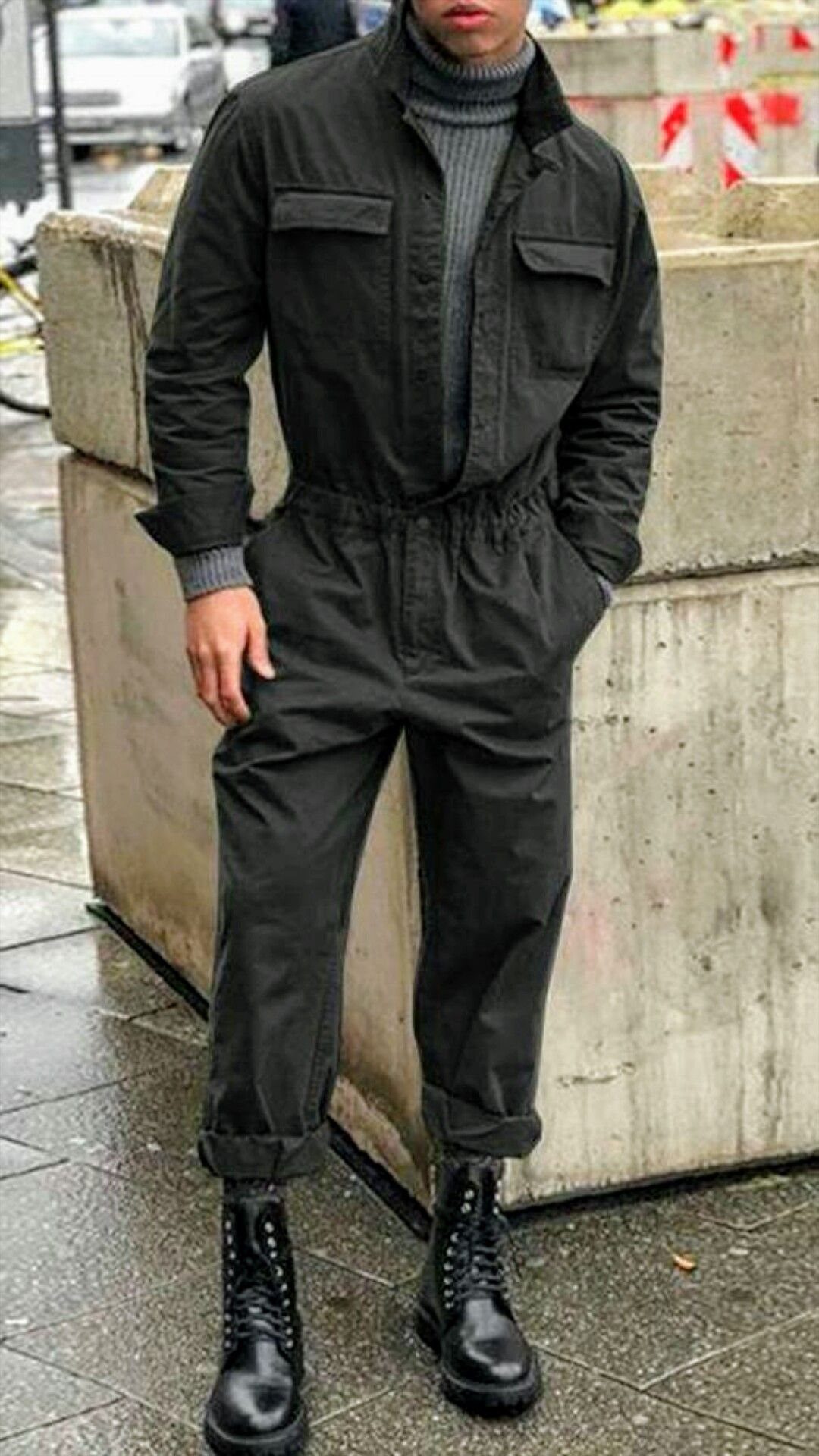 Man wearing a black jumpsuit, grey turtleneck, and black combat boots standing outdoors.