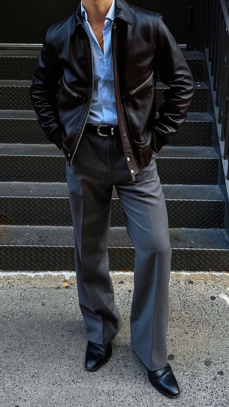 Man wearing a brown leather jacket, blue shirt, grey trousers, and black boots standing on stairs.