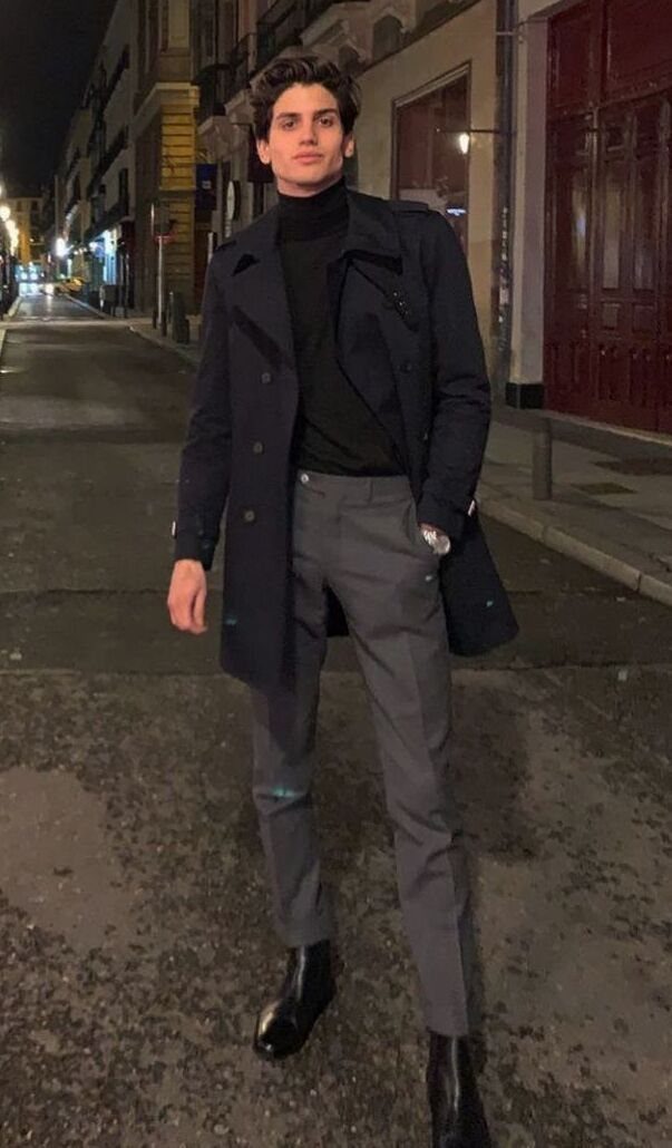 Young man posing in a navy trench coat, black turtleneck, and grey trousers on a city street at night.