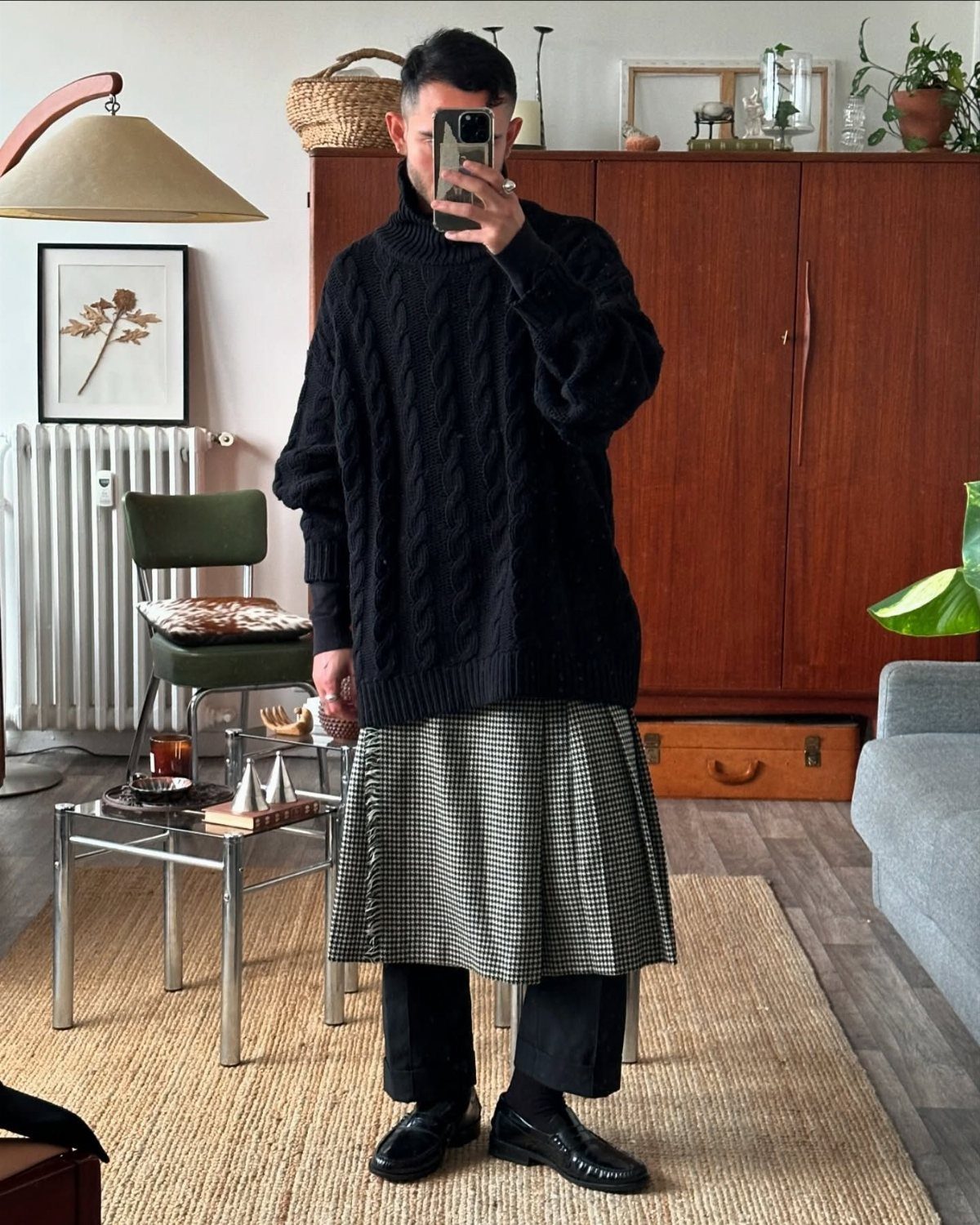 A person wearing a black cable-knit sweater and a grey plaid midi skirt standing indoors.