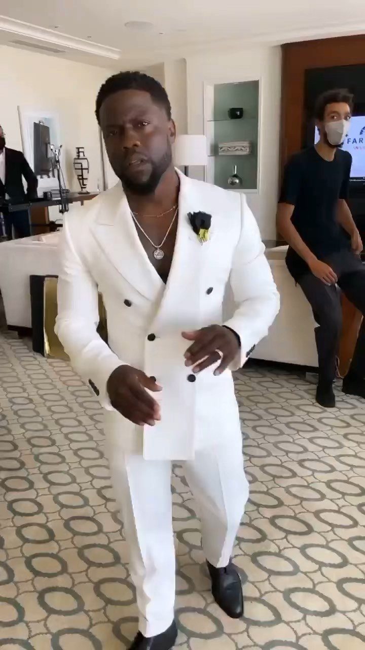 A man in a stylish white double-breasted suit and gold necklace gesturing in a room.