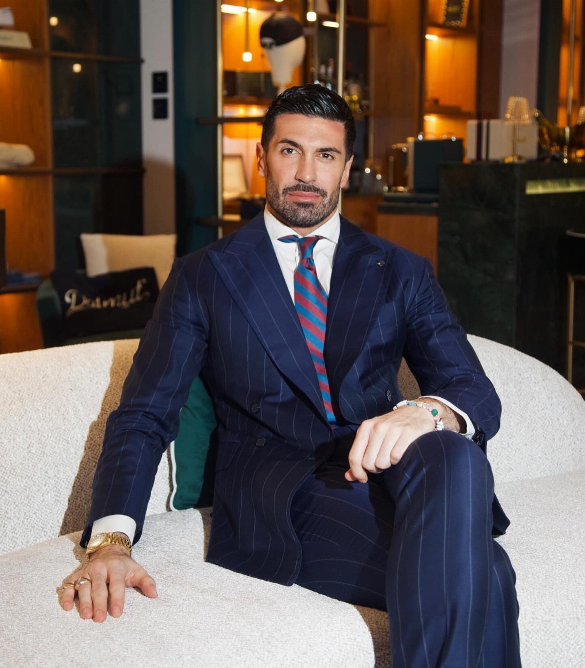 A man in a navy pinstripe suit and striped tie sitting on a white sofa.