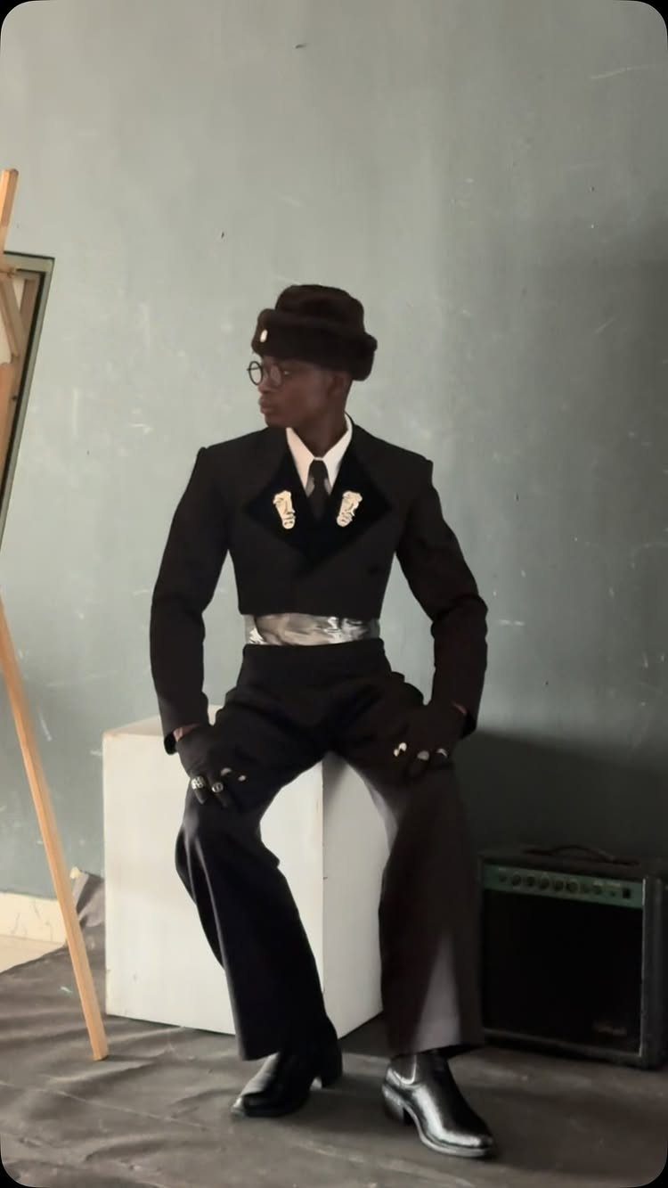 A man wearing an avant-garde black cropped jacket and wide-leg trousers sits on a white block.