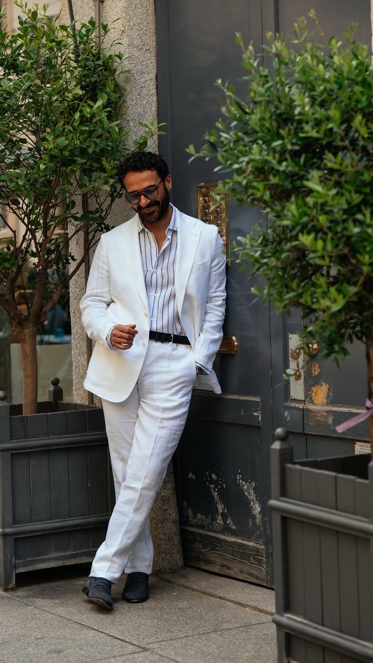 A man wearing a white linen suit and striped shirt leaning against a dark door.