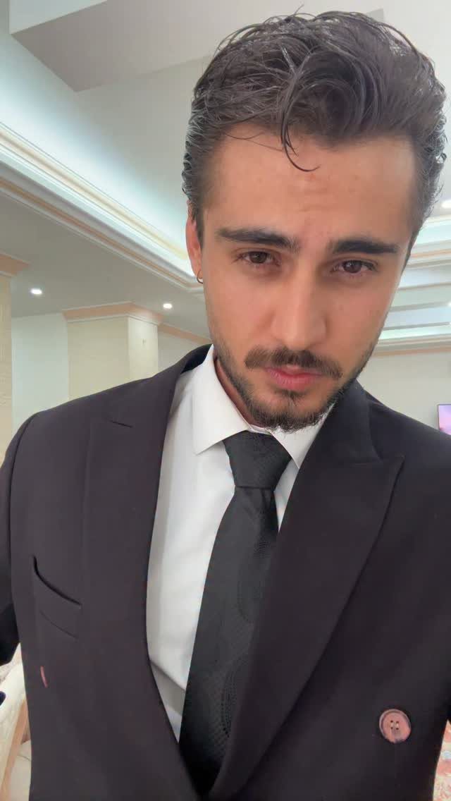 A man wearing a black suit, white shirt, and black tie looking at the camera.