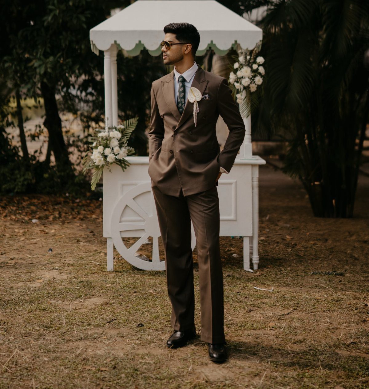 Man in a brown suit with a boutonniere and sunglasses standing by a white cart.