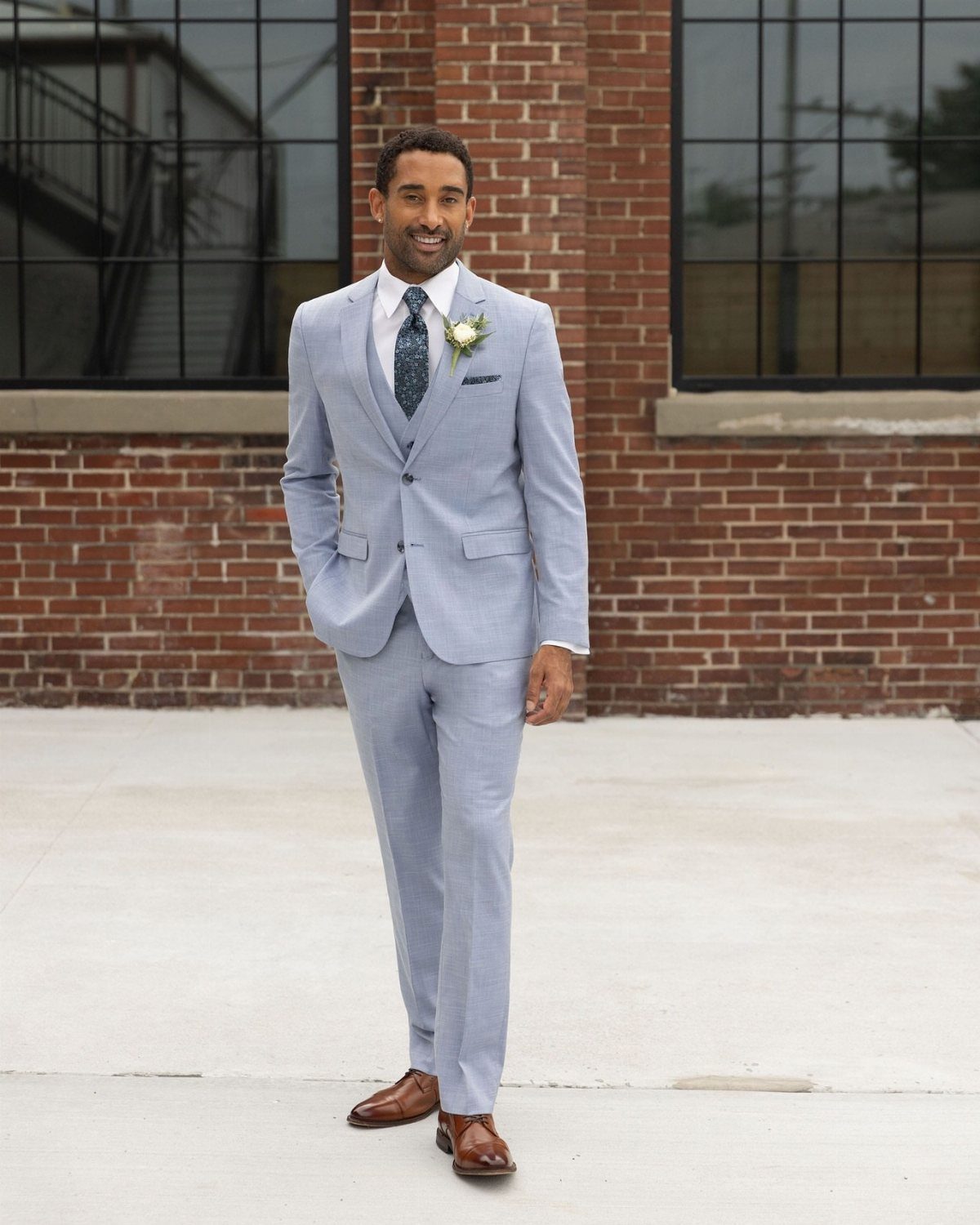 A man in a light blue three-piece suit and brown shoes standing against a brick wall.